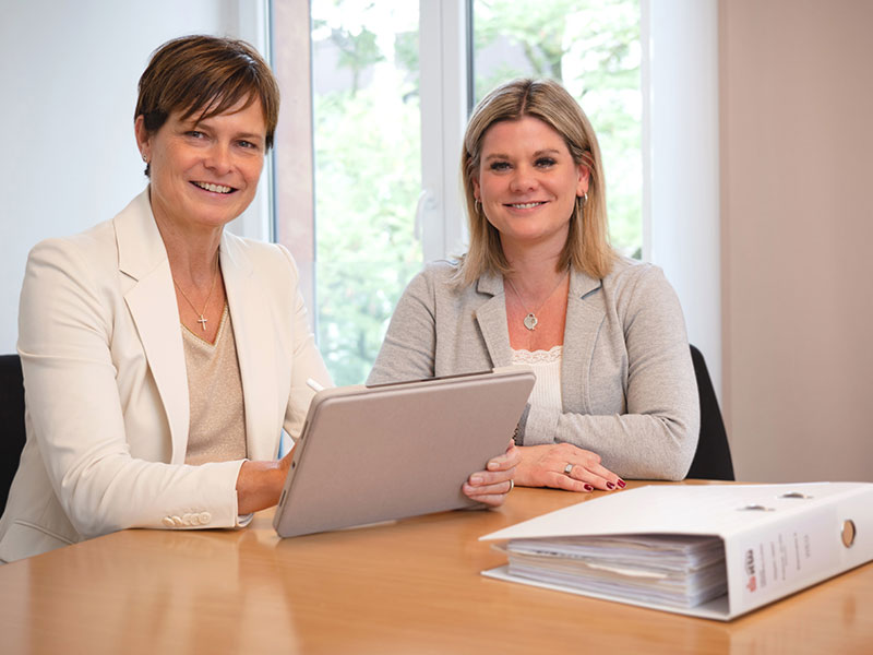 Katja Döring und Sarah Juraschek gemeinsam an einem Schreibtisch sitzend mit einem Tablet